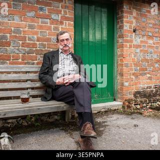 Elderly man with a mustache sitting on a wooden bench outside a rural pub in Oxfordshire Stock Photo