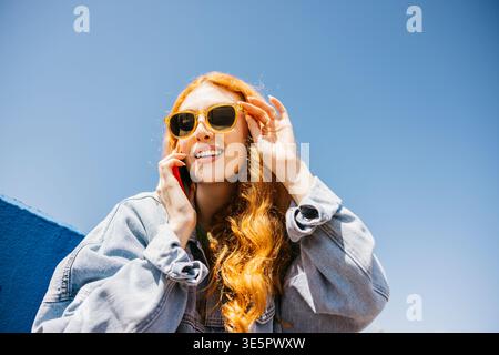 Redhead woman using a mobile phone, adjusting her sunglasses under a bright blue sky Stock Photo