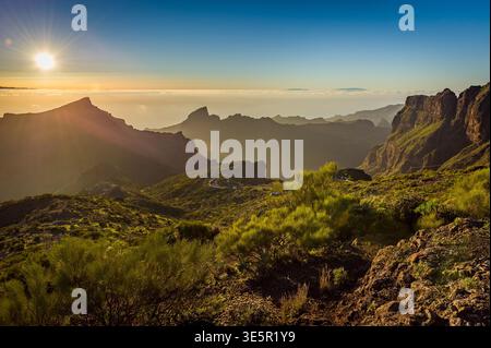 Sunlit Tenerife Mountains With Serpentine Road, Sharp Peaks, Soft Haze and Green Vegetation, Dramatic Landscape of Rugged Terrain and Deep Mountain Va Stock Photo