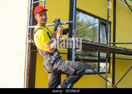 Construction worker sitting on scaffold with power tool, facade restoration, painting and installation, handyman service at home, professional repairs Stock Photo