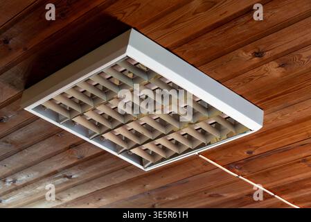 A fluorescent ceiling light fixture with a metallic grid is mounted on a wooden paneled ceiling, viewed from below in a rustic indoor setting. Stock Photo