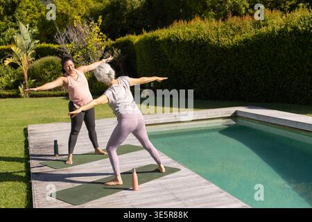 Diverse senior women practicing standing yoga poses on deck mats by pool in leggings with bottles Stock Photo