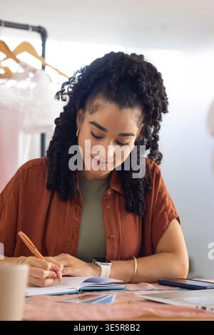 African American woman sitting at table writing in open notebook, wearing rust shirt, holding pen Stock Photo