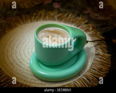 Hot cappuccino served in a pastel green designer cup placed on a rustic woven placemat, cozy still life composition with soft lighting Stock Photo