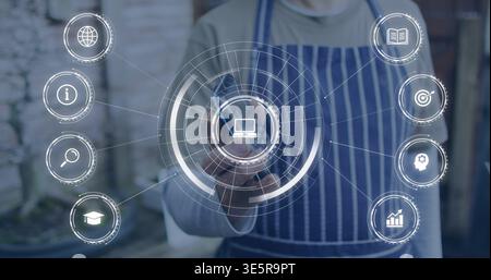 Pointing mature man in striped apron touching virtual circular interface with laptop icon at shop Stock Photo