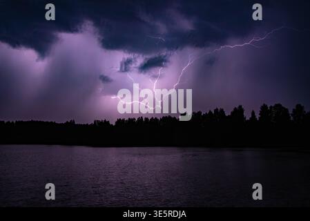 Powerful lightning bolts illuminate a dark stormy sky over a forest and calm lake at night. Stock Photo