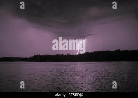 Lightning strikes over a dark sky above a lake, illuminating clouds and water with a purple glow. Stock Photo
