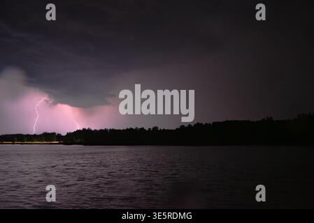 Intense lightning bolts illuminate dark storm clouds over a lake, casting a purple glow across the water and horizon. Stock Photo