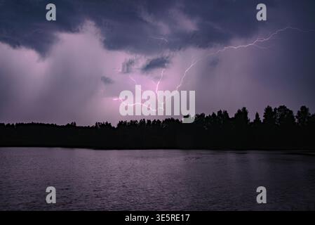 Powerful lightning bolts illuminate a dark stormy sky over a forest and calm lake at night. Stock Photo