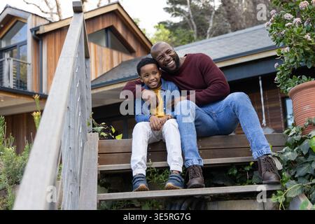 African American father and son sitting on wooden steps of wood-clad house, terracotta pot nearby Stock Photo