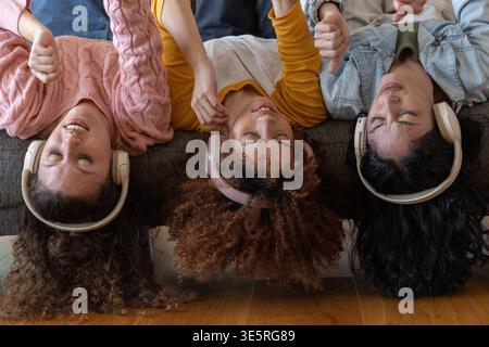 Diverse female friends lying upside down on sofa in living room wearing over-ear headphones Stock Photo