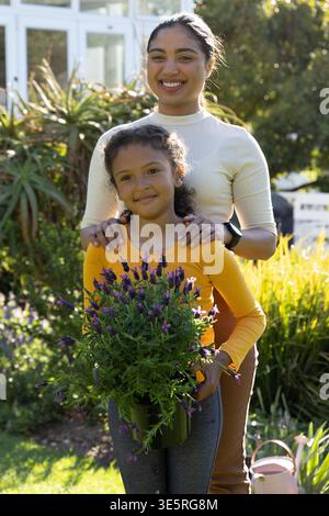 Mother and daughter standing in sunlit garden holding potted purple plant, pink watering can nearby Stock Photo