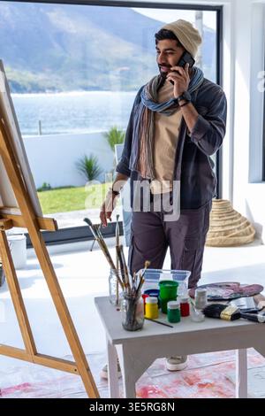Male artist in studio wearing beanie, standing by easel near window, phoning while reaching brushes Stock Photo