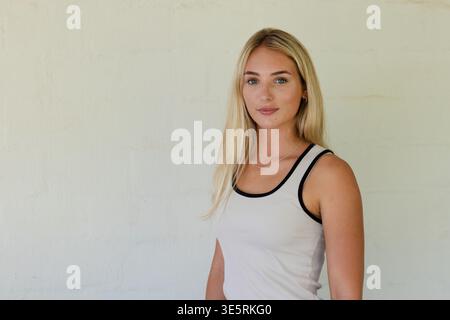 Woman standing by cream plaster wall wearing white tank with black trim, thin necklace, copy space Stock Photo