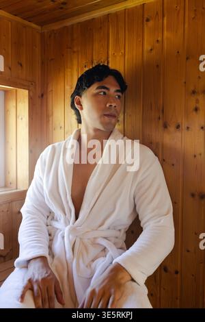 Man sitting on wooden bench in wood-paneled sauna cabin wearing white plush bathrobe, gazing right Stock Photo