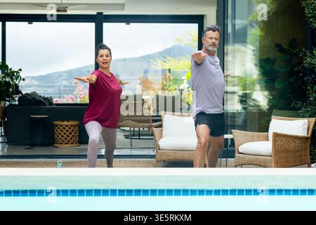 Senior couple practicing standing yoga in activewear on poolside deck with water bottle and plants Stock Photo