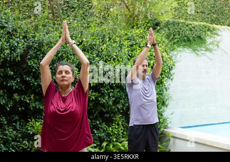 Mature couple practicing standing yoga pose in backyard garden by pool with wristwatches visible Stock Photo
