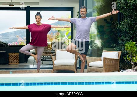 Diverse mature couple practicing tree pose in activewear balancing on patio near swimming pool Stock Photo