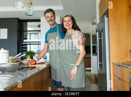 Diverse mature couple preparing salad at kitchen island wearing gray-green aprons with vegetables Stock Photo