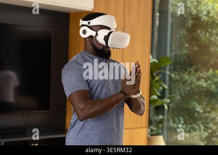 African man standing in living room wearing white VR headset, blue T-shirt, showing smartwatch Stock Photo