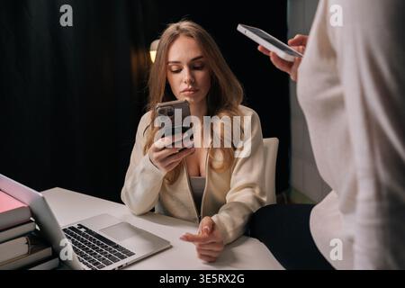 Two beautiful young female colleagues interacting with smartphones, one sitting at desk with laptop and books, other leaning on desk, both engaged in Stock Photo