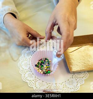 Close up of a woman hands pouring colorful sugar sprinkles into a pink paper cupcake liner. Stock Photo