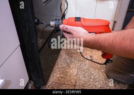 Close-up of a man drilling a hole in an oven door during a DIY repair, set in a realistic domestic kitchen environment. Stock Photo