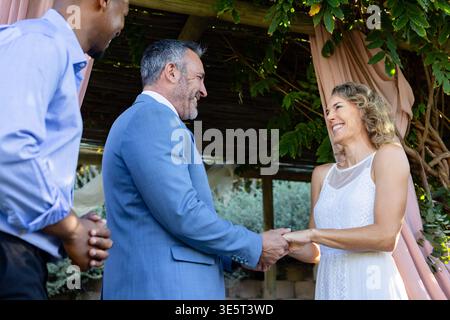Officiant asking vows, groom and bride holding hands under arbor exchanging rings, marking wedding Stock Photo