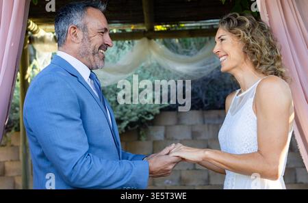 Couple standing under pergola in garden holding hands wearing blue suit and sleeveless lace dress Stock Photo