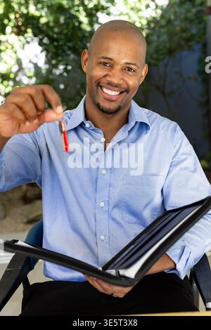 Adult African man smiling, wearing blue shirt and black pants, holding red key, portfolio on patio Stock Photo