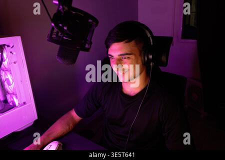 A young man wearing a black shirt and headphones smiles while looking at his computer monitor. The room features a gaming setup with a microphone and Stock Photo