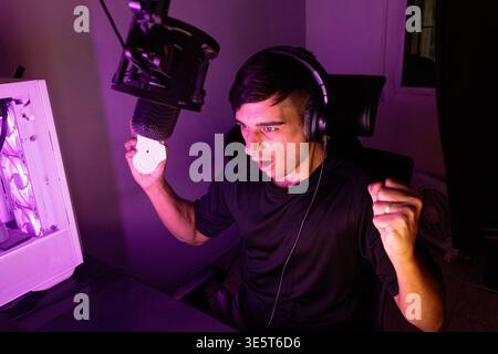 A young caucasian man with headphones shows an expression of joy and celebration with his mouth open and arms raised while sitting at his computer. Th Stock Photo