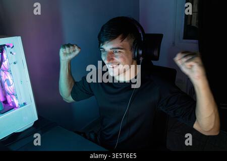 A joyful young caucasian man raises his fists in celebration while looking at his computer screen. The room is illuminated by cool blue neon lights fr Stock Photo