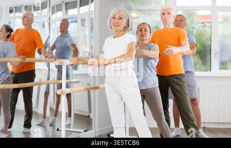Active mature woman practicing battement tendu holding by ballet barre together with others Stock Photo