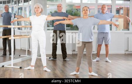 Experienced mature female ballet dancer, dressed in sportswear executing dance moves with focused precision among group of aged people rehearsing in Stock Photo