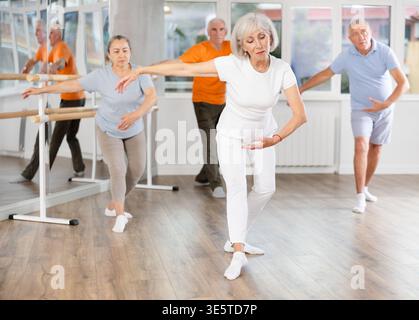 Happy aged woman in sportswear confidently leading ballet class with dancers following movements in dance studio Stock Photo