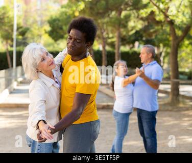 Carefree senior multiracial couples dancing in pairs outdoors during sunny day at park Stock Photo