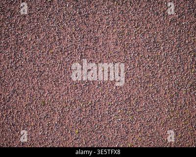 detailed shot of a textured reddish-brown render wall or running track surface. The grainy composition features small, multi-colored pebbles and crush Stock Photo