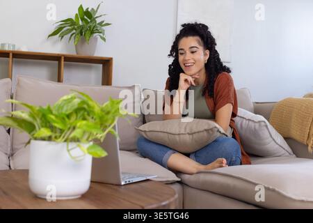 Adult woman sitting crosslegged holding pillow leaning toward laptop, typing at home, copy space Stock Photo