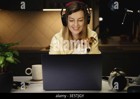 Happy smiling woman sitting at table with her dog and looking in laptop screen. Female freelancer working from home office at night with pet Stock Photo
