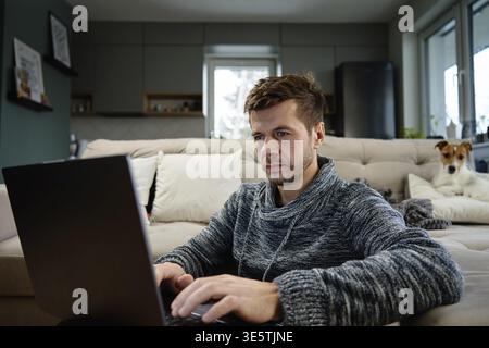 Man using laptop in living room, dog lying on sofa at background. Freelancer working at home workplace. Remote work and work from home concept Stock Photo