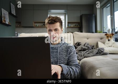 Man using laptop in living room, dog lying on sofa at background. Freelancer working at home workplace. Remote work and work from home concept Stock Photo