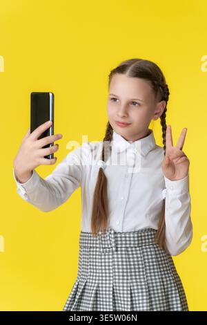 Happy schoolgirl takes selfie. She wears school uniform and makes peace sign. Bright yellow background provides copy space. Modern concept of youth, t Stock Photo