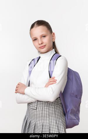 Portrait of preteen student with confident look. Schoolgirl wears uniform and purple backpack, arms crossed. High key studio shot on white background. Stock Photo