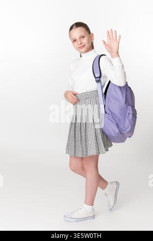 Full length studio portrait of happy preteen schoolgirl ready for school. Girl wears uniform, carries purple backpack, and waving gesture with smile. Stock Photo