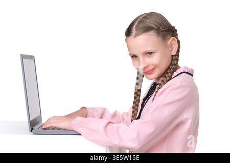 Portrait of happy preteen girl with two braids in pink shirt studying online. Schoolgirl types on her computer, looking at the camera with smile. Clea Stock Photo