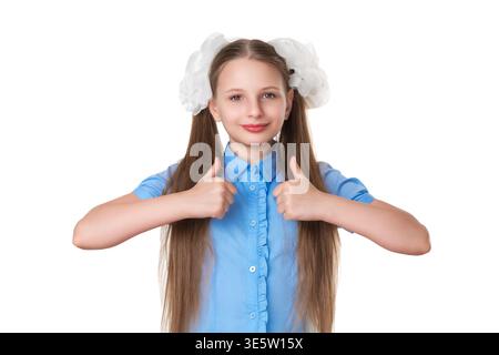 Happy student girl with long hair in pigtails and big white bows gives thumbs up gesture. Positive child expressing approval and success. Studio portr Stock Photo