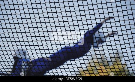 Person jumping on trampoline seen through safety net against blue sky Stock Photo