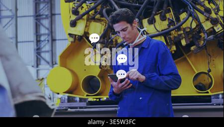 Tapping tech in blue coverall checking phone at factory, with yellow machine and overlays Stock Photo