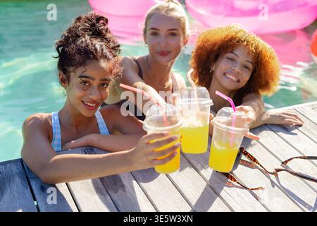 Diverse female friends in swimsuits leaning on wooden poolside deck, holding clear plastic cups Stock Photo